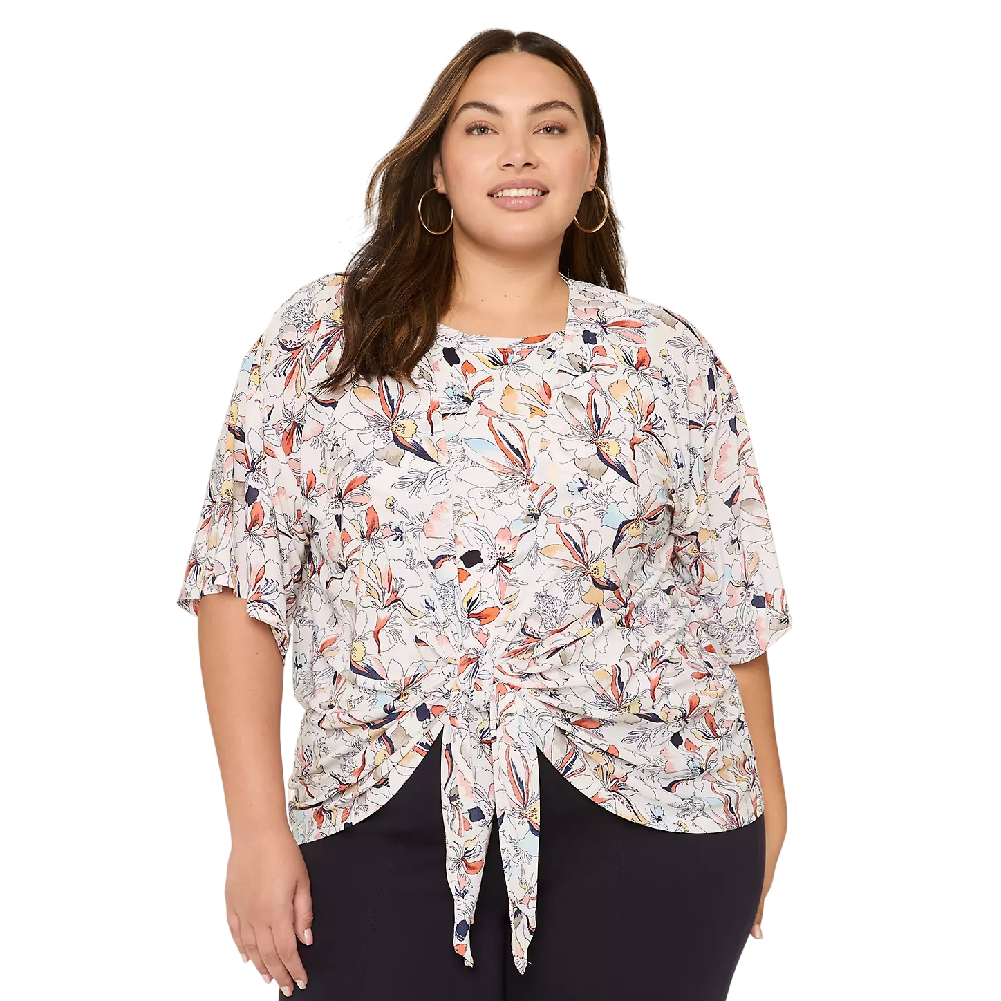 A woman is wearing a patterned blouse with short sleeves and a front-tie design, paired with dark pants. The blouse features a colorful floral design in shades of orange, blue, and pink.