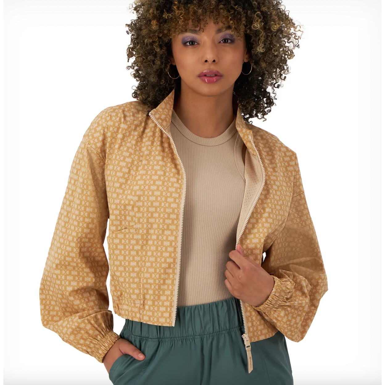 A woman is wearing a yellow patterned Champion jacket with a zip closure and a ribbed collar, paired with a beige top and green pants.
