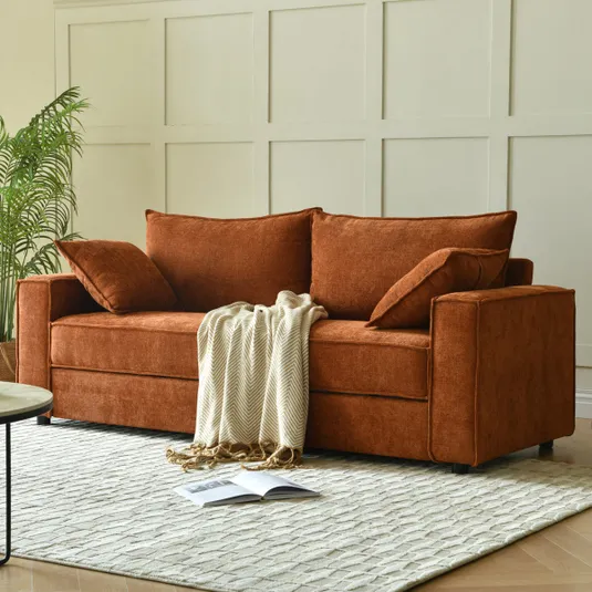 A rust-colored sofa with cushions is adorned with a cream throw blanket. It sits on a textured light beige rug, with a plant and a wooden wall paneling in the background, alongside an open magazine on the floor.
