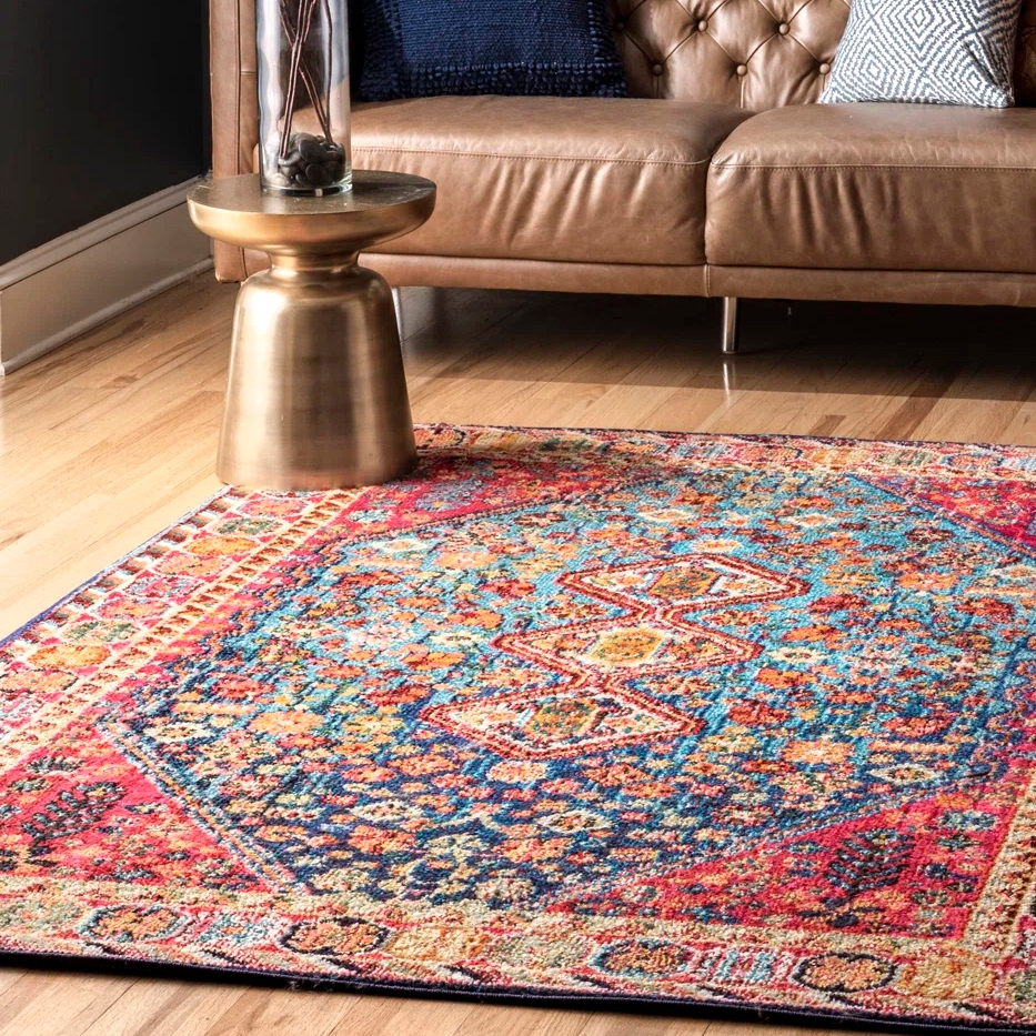 A colorful Bohemian area rug with a complex pattern featuring blue, red, and orange tones lies on a wooden floor. It is situated in front of a leather sofa and next to a gold-toned side table.