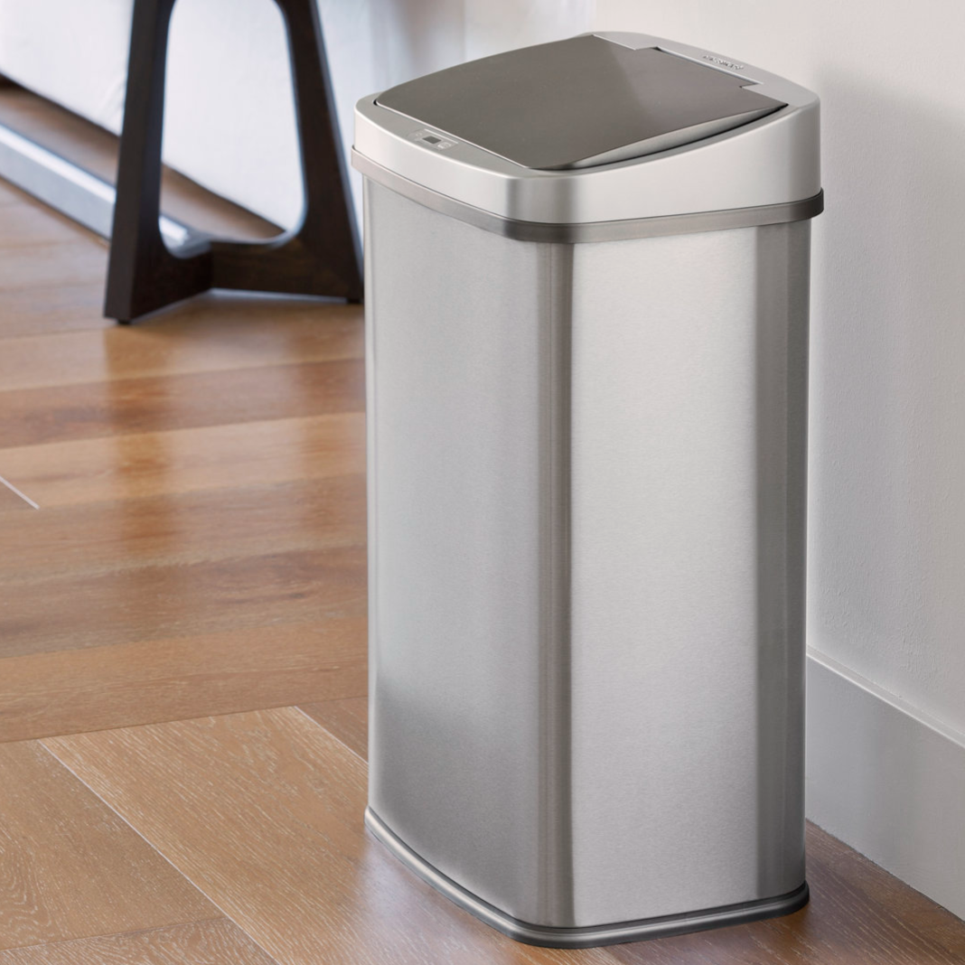 A sleek, stainless steel trash can with a sensor-operated lid, designed for hands-free operation. It's positioned in a modern interior setting with wooden flooring.