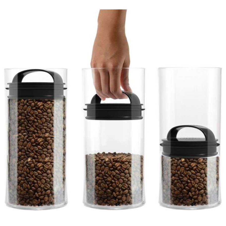Three cylindrical airtight containers in a row with black lids, filled with coffee beans; one lid being lifted by a hand.