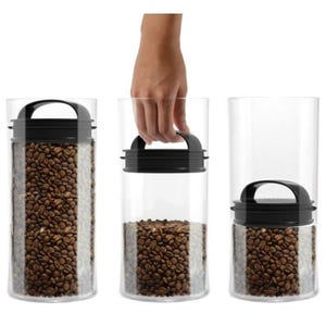 Three cylindrical airtight containers in a row with black lids, filled with coffee beans; one lid being lifted by a hand.