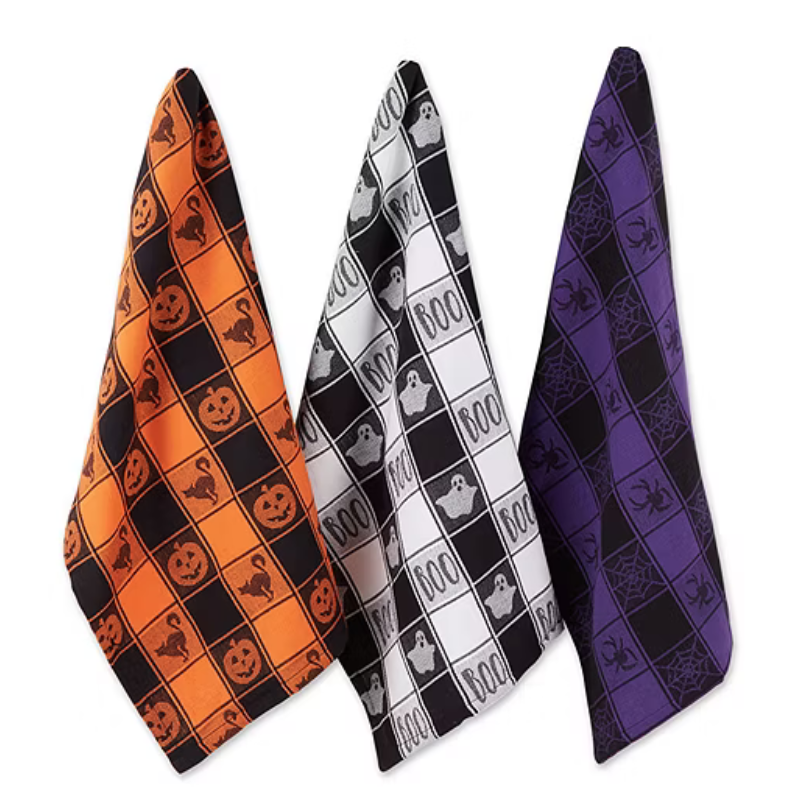 Three Halloween-themed kitchen towels feature designs with pumpkins and cats on orange, ghosts and \“BOO\“ on black and white, and spiders and webs on purple.
