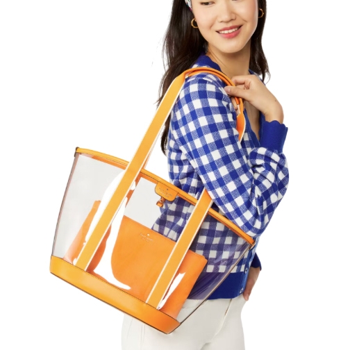 A woman carries a transparent tote bag with orange accents and handles. She wears a blue and white checkered sweater and white pants.