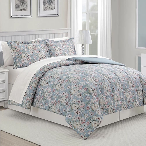 A floral patterned bedding set includes a comforter, bed skirt, and matching pillow shams, displayed on a made bed in a bright room with a lamp and framed wall art.