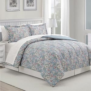 A floral patterned bedding set includes a comforter, bed skirt, and matching pillow shams, displayed on a made bed in a bright room with a lamp and framed wall art.