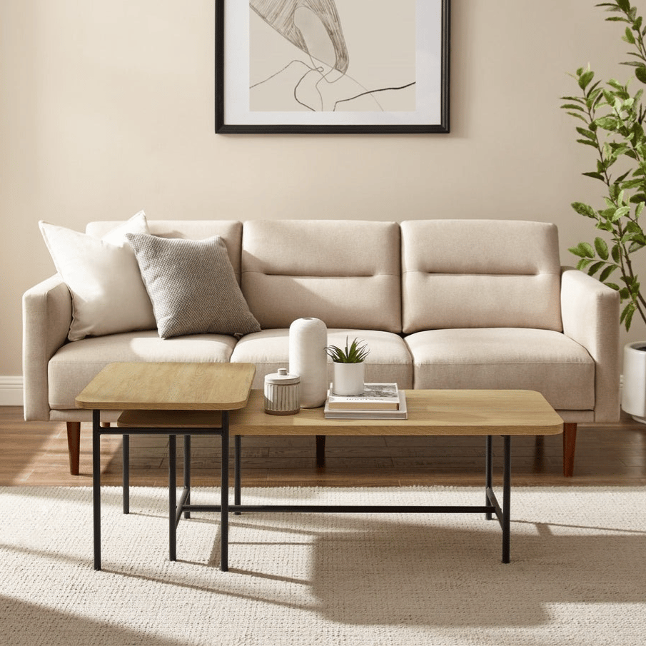 A beige sofa with a tufted design is accompanied by a dual-level wooden coffee table with metal legs, set on a textured rug. A minimalist abstract art piece is framed above, and a tall potted plant adds greenery to the room.