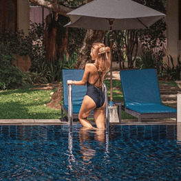 A person in a black swimsuit stands in a clear blue swimming pool, next to blue lounge chairs and a gray umbrella, surrounded by greenery.