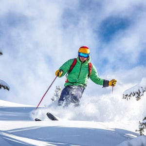A skier wearing a green jacket, yellow gloves, red helmet, and blue goggles skis downhill on fresh powder, holding ski poles.