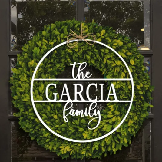 A decorative green wreath with a white overlay displays the text \“The Garcia Family,\“ hanging on a glass-paneled door, tied with twine at the top.