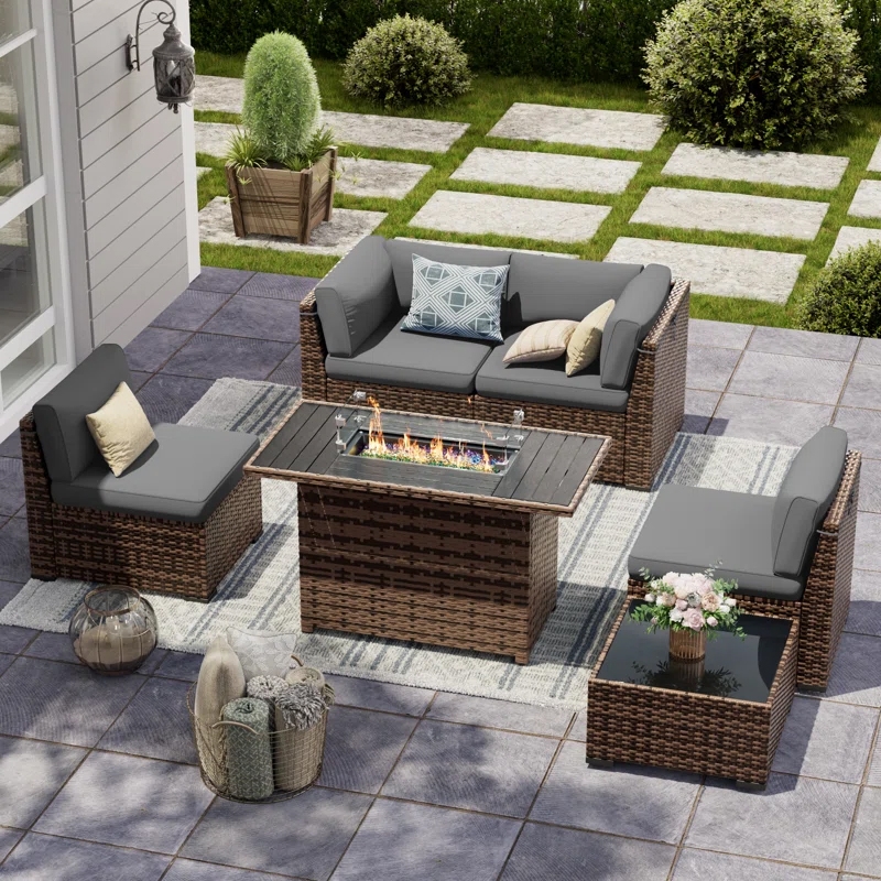 Outdoor patio set with gray cushions, rattan-style loveseat, two chairs, and side table. Features a rectangular fire pit table with flames and decorative elements like a rug, a lantern, and potted plants.