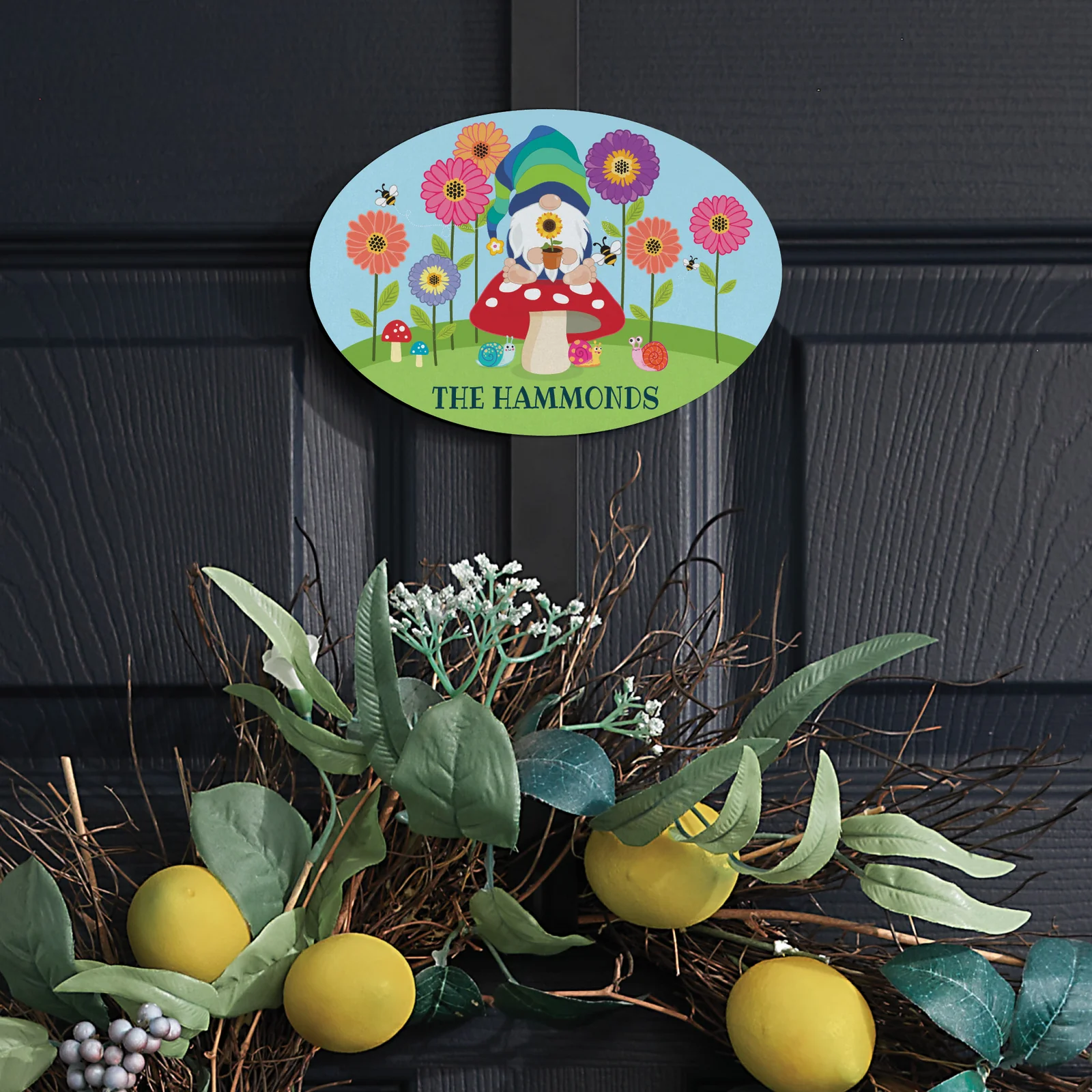 A decorative door sign features a whimsical gnome sitting on a mushroom with colorful flowers and bees, personalized with \“The Hammonds.\“ Below, a wreath with lemons and greenery decorates the door.