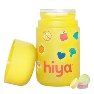 A yellow container labeled \“hiya\“ features colorful stickers of fruits and vegetables, with three assorted-color chewable tablets next to it.