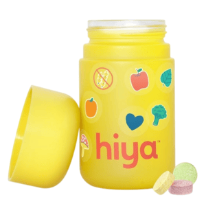 A yellow container labeled \“hiya\“ features colorful stickers of fruits and vegetables, with three assorted-color chewable tablets next to it.