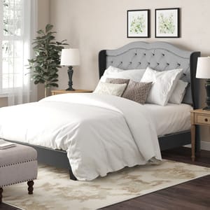 A bedroom features a gray tufted headboard bed with white bedding, side tables with lamps, a bench, and a plant, all set on a patterned rug. Two framed artworks are hung above the bed.