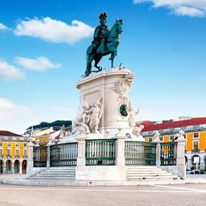 A statue depicting a rider on horseback is prominently displayed, surrounded by elaborate sculptures. The structure is set in a plaza with colorful historic buildings in the background.