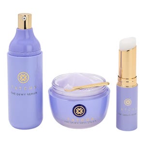 A 3-piece Tatcha skincare set includes The Dewy Serum in a tall bottle, The Dewy Skin Cream in a jar with a small spatula, and The Serum Stick in a tube. All are in light purple packaging with gold accents.