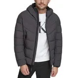 A man is wearing a dark gray puffer jacket with a hood over a white sweatshirt featuring a large \“K\“ and the brand name \“Klein Jeans.\“