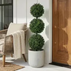 A potted topiary with three spherical sections stands next to a wicker chair draped with a beige knitted throw, set against a white wall and a wooden door.
