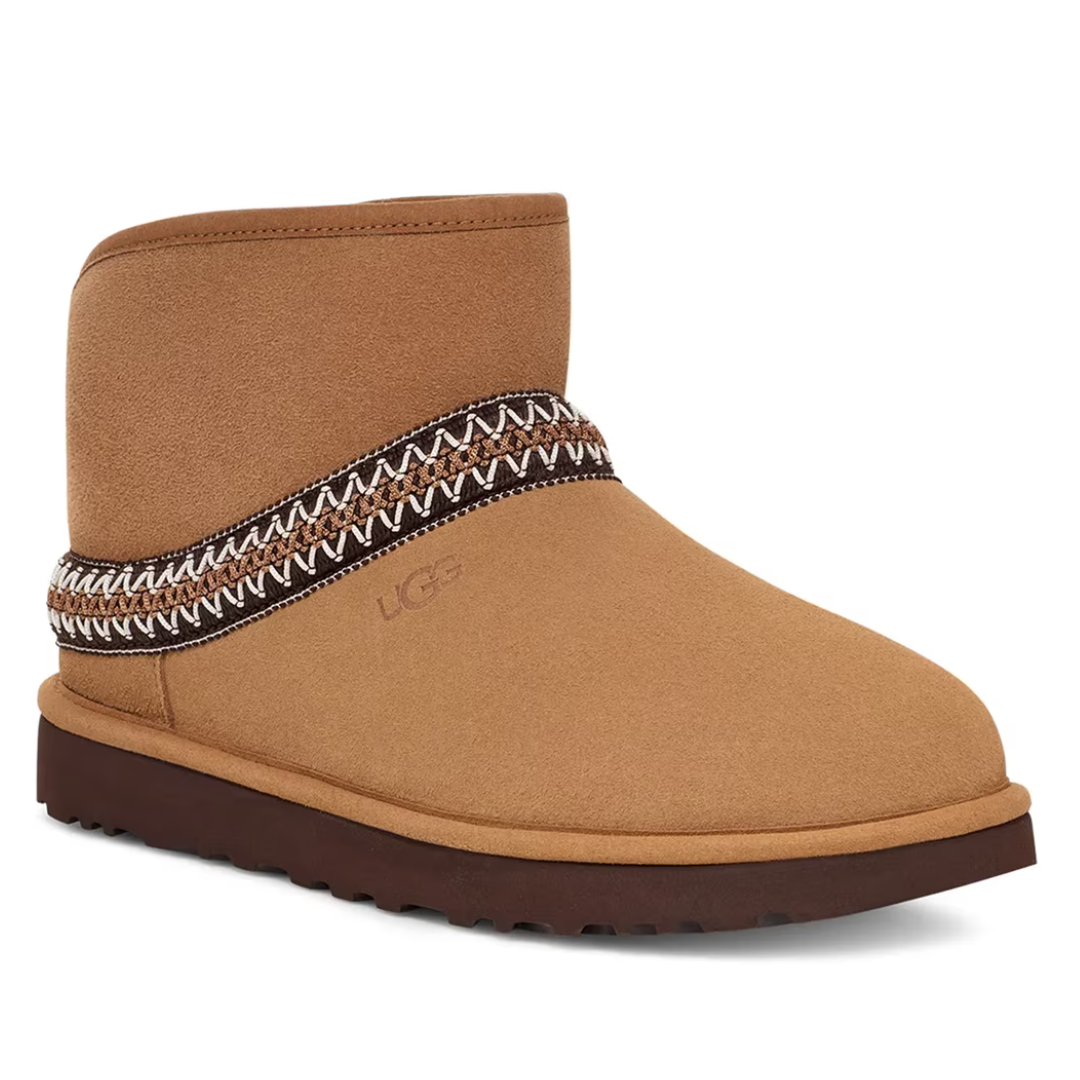 A tan boot with a patterned trim, featuring a short shaft and a treaded sole, displaying the UGG logo on the side.
