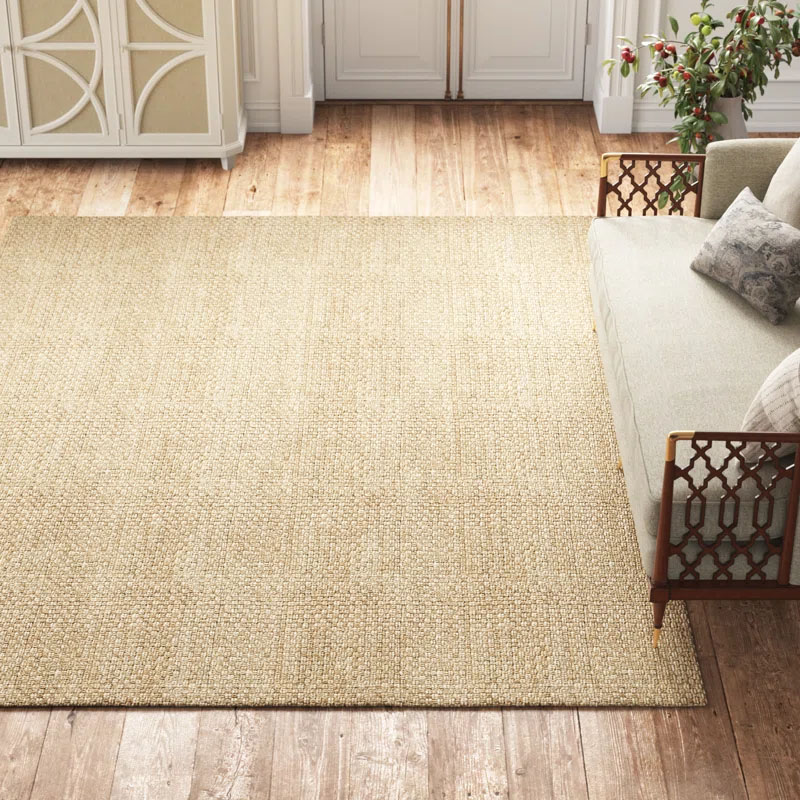 Kelly Clarkson's 5' x 8' Soprano Jute Rug is a textured, natural-colored rug placed on a wooden floor beside a patterned sofa and decorative pillows, offering an elegant and cozy look.
