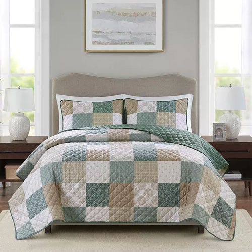 A neatly made bed features a patchwork quilt with matching pillow shams, blending soft greens, beiges, and whites in a checkerboard pattern, set between two bedside tables with lamps and a framed artwork above the headboard.