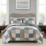 A neatly made bed features a patchwork quilt with matching pillow shams, blending soft greens, beiges, and whites in a checkerboard pattern, set between two bedside tables with lamps and a framed artwork above the headboard.