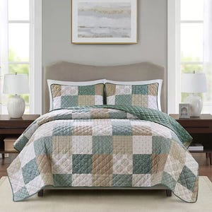 A neatly made bed features a patchwork quilt with matching pillow shams, blending soft greens, beiges, and whites in a checkerboard pattern, set between two bedside tables with lamps and a framed artwork above the headboard.