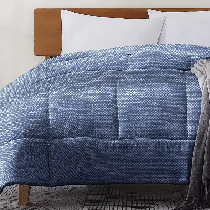 A blue textured comforter is on a bed with a wooden headboard, accompanied by white pillows and a gray blanket draped over the edge.