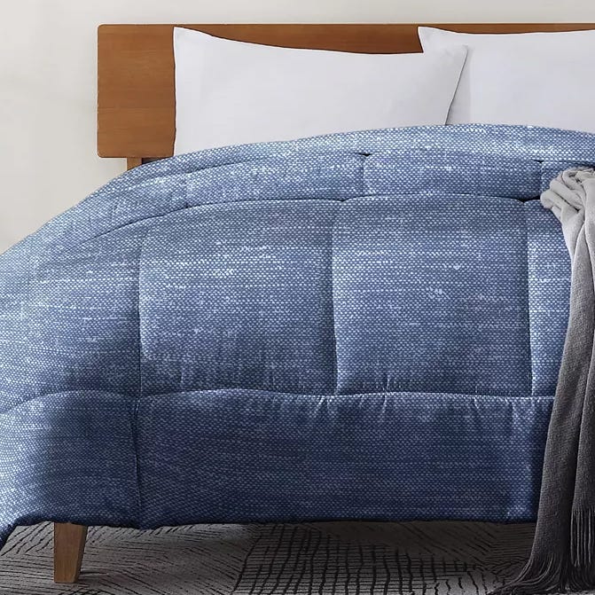A blue textured comforter is on a bed with a wooden headboard, accompanied by white pillows and a gray blanket draped over the edge.