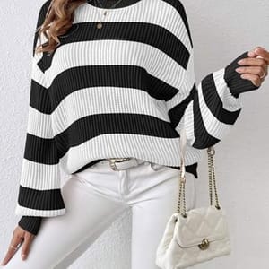 A woman is wearing a black and white striped sweater with a round neckline and long sleeves, paired with a white belt and white pants.