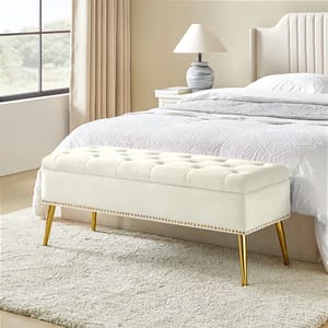 A cream-colored, tufted flip-top storage bench with gold legs, positioned at the foot of a bed in a modern bedroom setting.