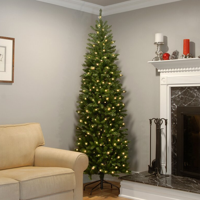 A tall, slender, pre-lit artificial Christmas tree with white lights stands in a living room corner near a fireplace and an armchair.