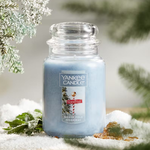 Yankee Candle jar labeled \“North Pole Hideaway\“ featuring a light blue hue, surrounded by snow with pine branches in the background.