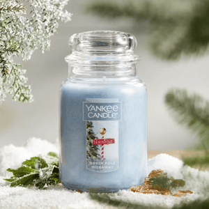 Yankee Candle jar labeled \“North Pole Hideaway\“ featuring a light blue hue, surrounded by snow with pine branches in the background.