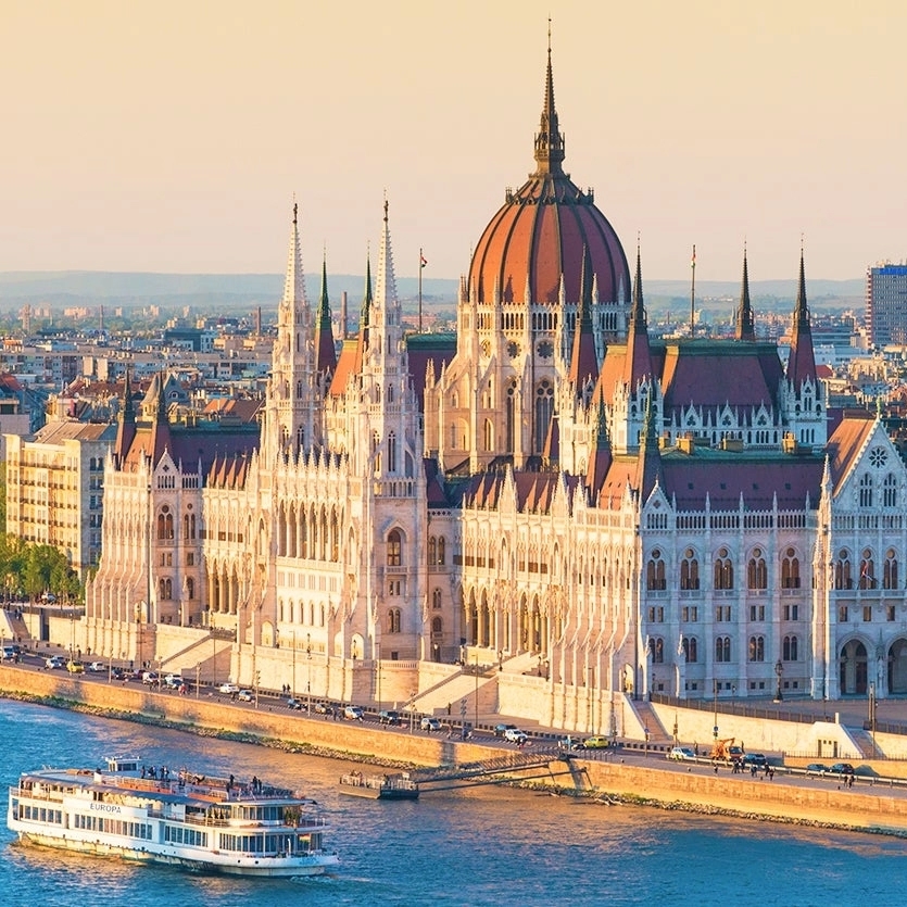The Hungarian Parliament Building is depicted on the bank of the Danube River with a cruise boat passing by.
