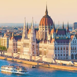 The Hungarian Parliament Building is depicted on the bank of the Danube River with a cruise boat passing by.