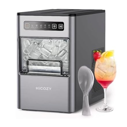 A portable ice maker with a silver exterior next to a spoon and a cocktail glass filled with a mixed drink and ice.