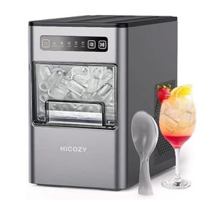 A portable ice maker with a silver exterior next to a spoon and a cocktail glass filled with a mixed drink and ice.