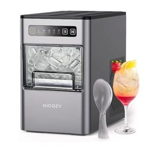 A portable ice maker with a silver exterior next to a spoon and a cocktail glass filled with a mixed drink and ice.