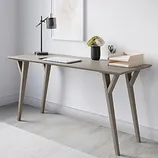 A minimalist desk setup featuring a slender wooden desk with angled legs, a black desk lamp, a small plant, a document organizer with papers, and scissors. A framed abstract art piece hangs on the wall above.