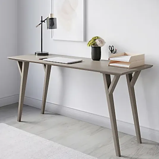 A minimalist desk setup featuring a slender wooden desk with angled legs, a black desk lamp, a small plant, a document organizer with papers, and scissors. A framed abstract art piece hangs on the wall above.