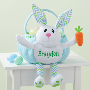 A stuffed bunny Easter basket with a carrot in one paw, featuring the name \“Brayden,\“ includes pastel-colored eggs inside. The bunny has zigzag-patterned ears and legs.