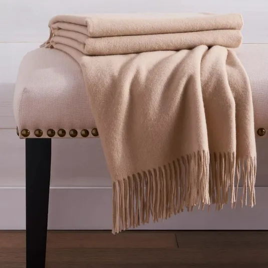 A folded beige blanket with fringe detailing is draped over a cushioned bench with nailhead trim and dark wooden legs.