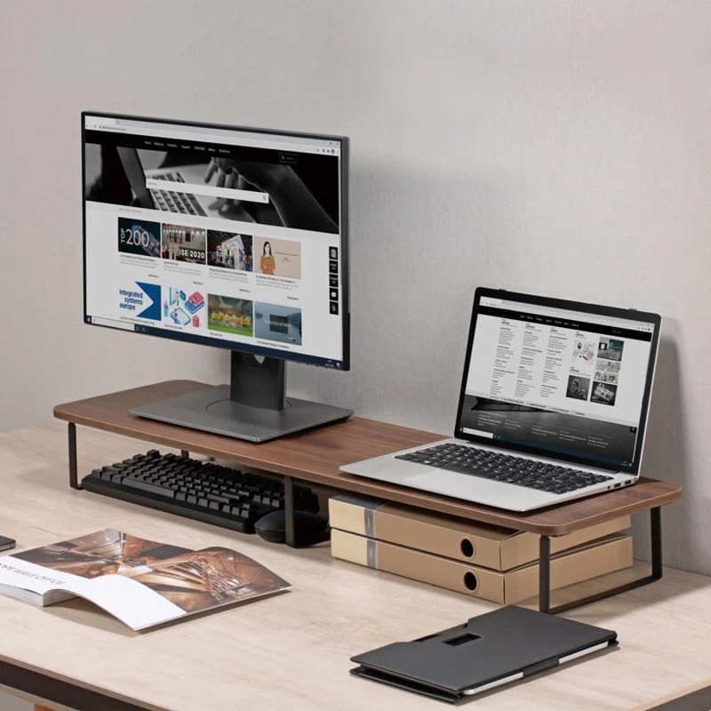 A desktop setup with a wooden monitor stand holds a computer monitor and a laptop. A keyboard is stored underneath the stand, and there are two document boxes stacked next to the laptop. A magazine and a closed folio or tablet are on the desk.