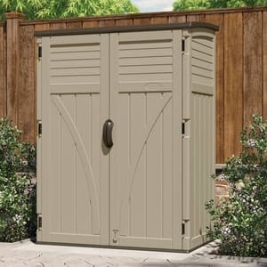The image shows a beige, vertical Suncast tool shed with double doors and a black handle, set against a wooden fence. It's surrounded by greenery, providing outdoor storage in a compact space.