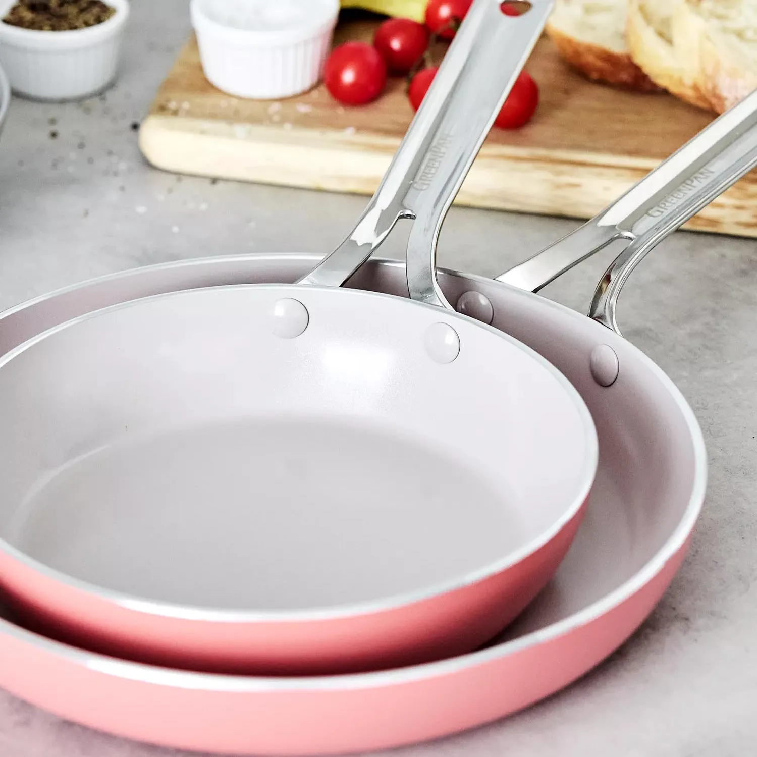 Two pink ceramic-coated frying pans with stainless steel handles, stacked on a kitchen counter. In the background, ingredients like cherry tomatoes and bread are visible on a cutting board.