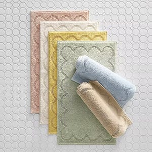 Five pastel-colored bath rugs in pink, white, yellow, green, and blue are laid on a hexagonal tile floor. They feature a textured scalloped design along the edges.