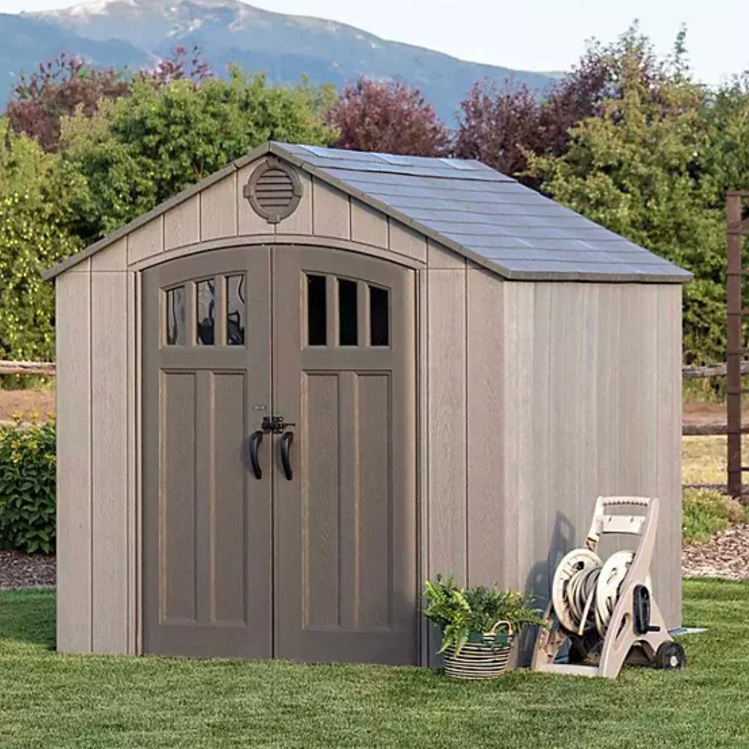 Suncast 8' Storage Shed features a double-door design with arched windows, shingle-style roof, and neutral color. A garden hose reel and potted plant are placed nearby, highlighting its use in a backyard setting.
